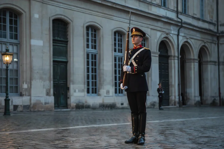 Un soldat en uniforme militaire de cérémonie est debout en poste sur une cour pavée, devant une façade de château aux arcades classiques.