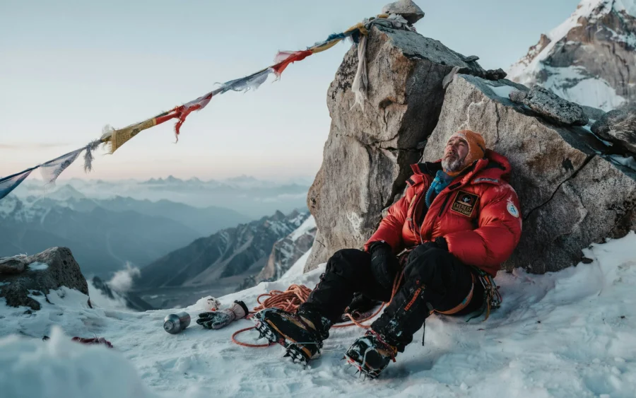 Alpiniste en combinaison rouge assis au sommet d'une montagne enneigée à côté de drapeaux de prière