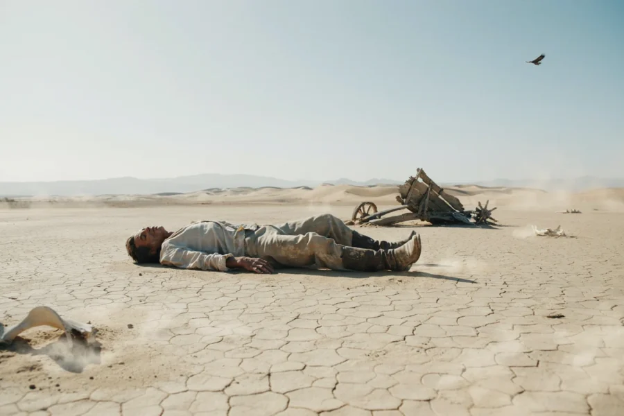 Un homme repose au sol dans un environnement dunal crackle sous un ciel bleu, tandis qu'un oiseau noir vole au-dessus.