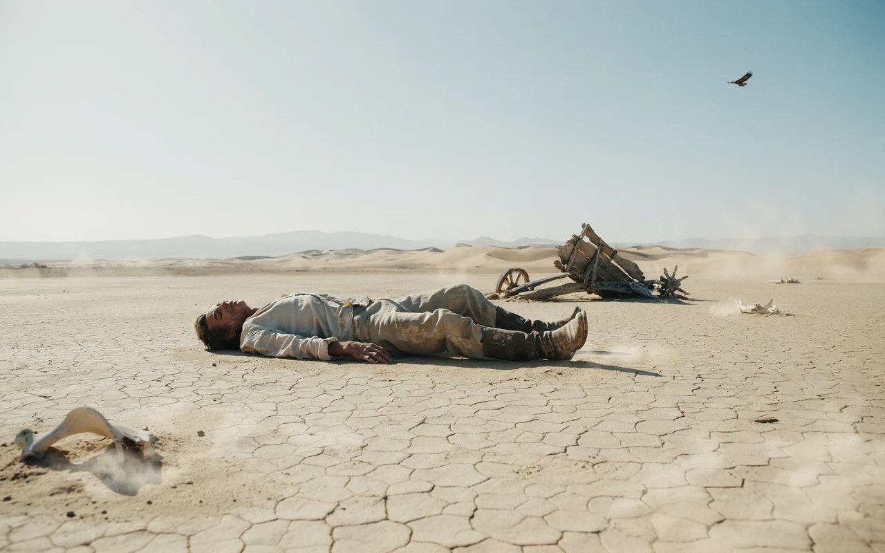 Un homme repose au sol dans un environnement dunal crackle sous un ciel bleu, tandis qu'un oiseau noir vole au-dessus.