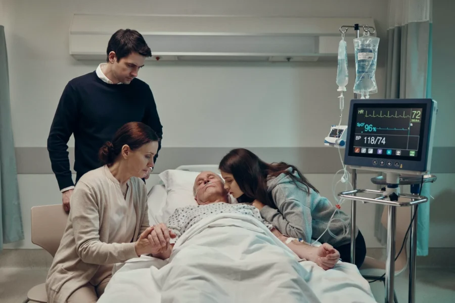 Une personne est allongée dans un lit d'hôpital entourée de trois proches tandis qu'un moniteur médical et des perfusions sont visibles.