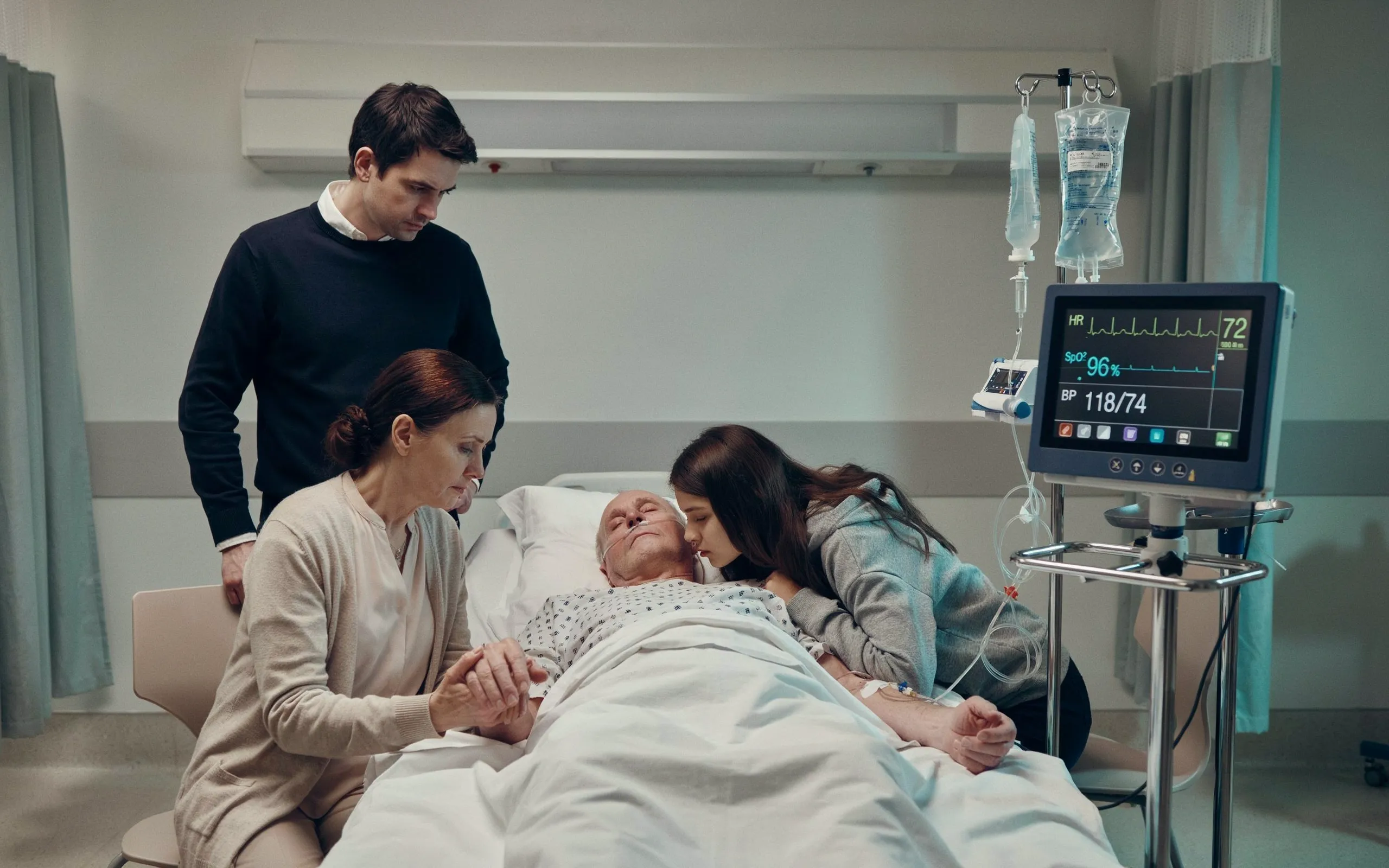 Une personne est allongée dans un lit d'hôpital entourée de trois proches tandis qu'un moniteur médical et des perfusions sont visibles.