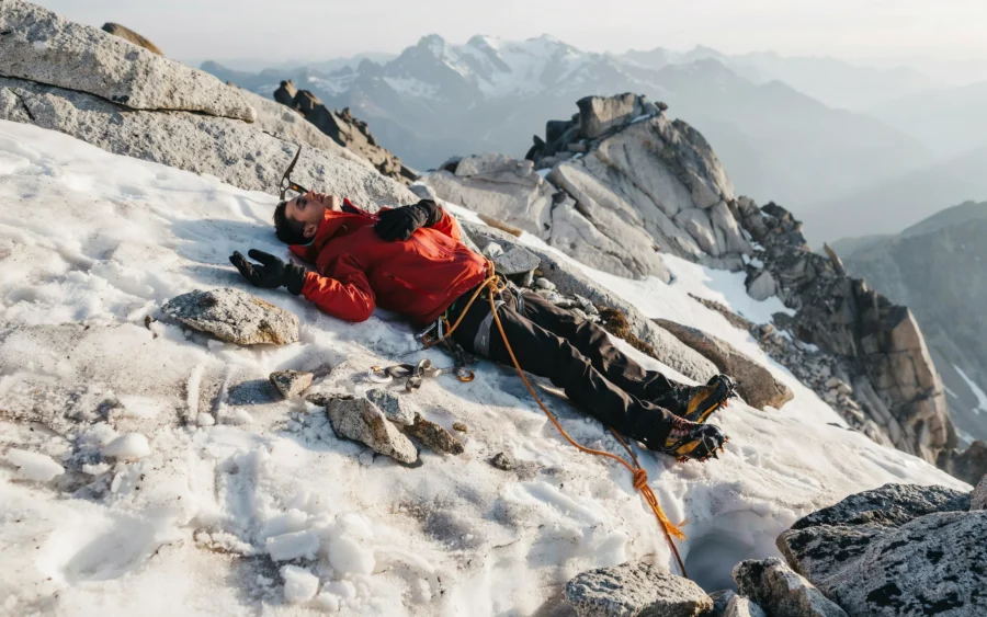 Des grimpeurs équipés progressent sur une pente rocheuse et enneigée, reliés par une corde orange. Le décor montagneux brumeux souligne l’altitude et les conditions extrêmes.