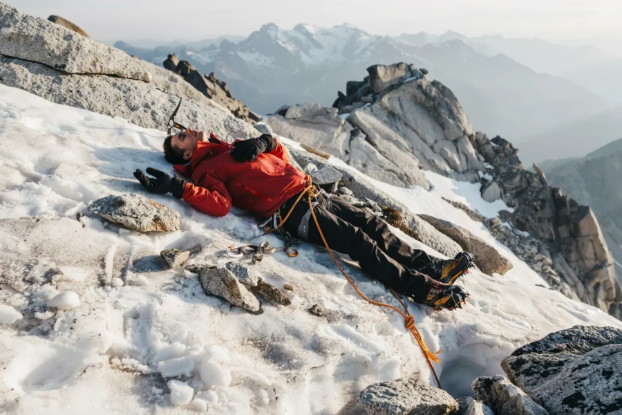 Des grimpeurs équipés progressent sur une pente rocheuse et enneigée, reliés par une corde orange. Le décor montagneux brumeux souligne l’altitude et les conditions extrêmes.