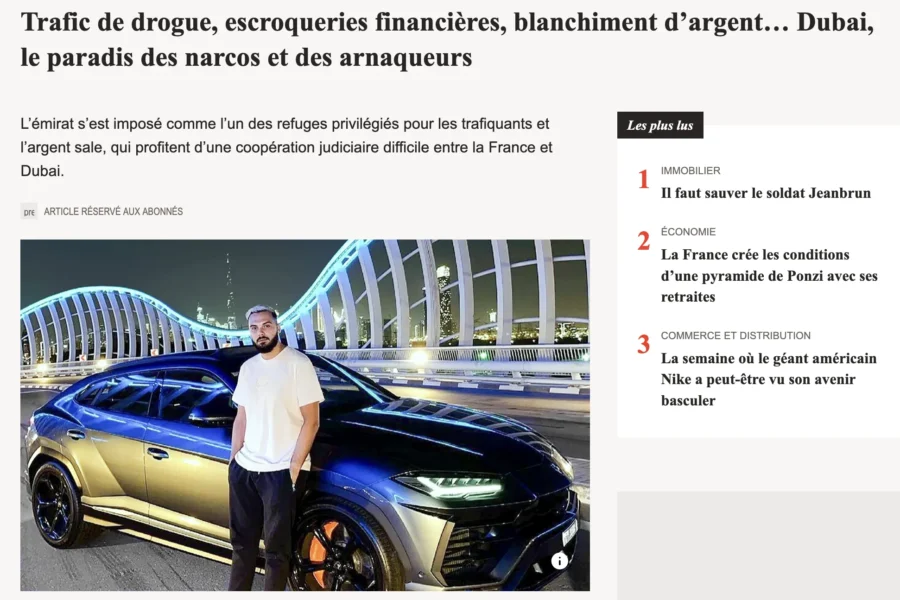 Un homme en chemise blanche se tient debout devant une automobile noire et or de grande taille sur un quai urbain.