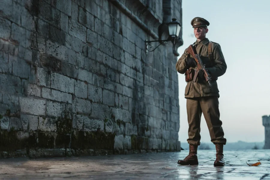 Un homme en uniforme militaire debout près d’un mur en pierre, tenant une arme, dans un cadre extérieur éclairé par une lumière douce.