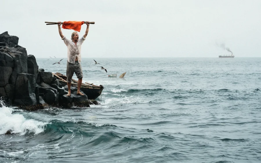 Un homme debout sur des rochers marins agite un linge orange sur une hampe fixée à une échelle.