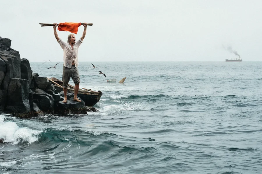 Un homme debout sur des rochers marins agite un linge orange sur une hampe fixée à une échelle.