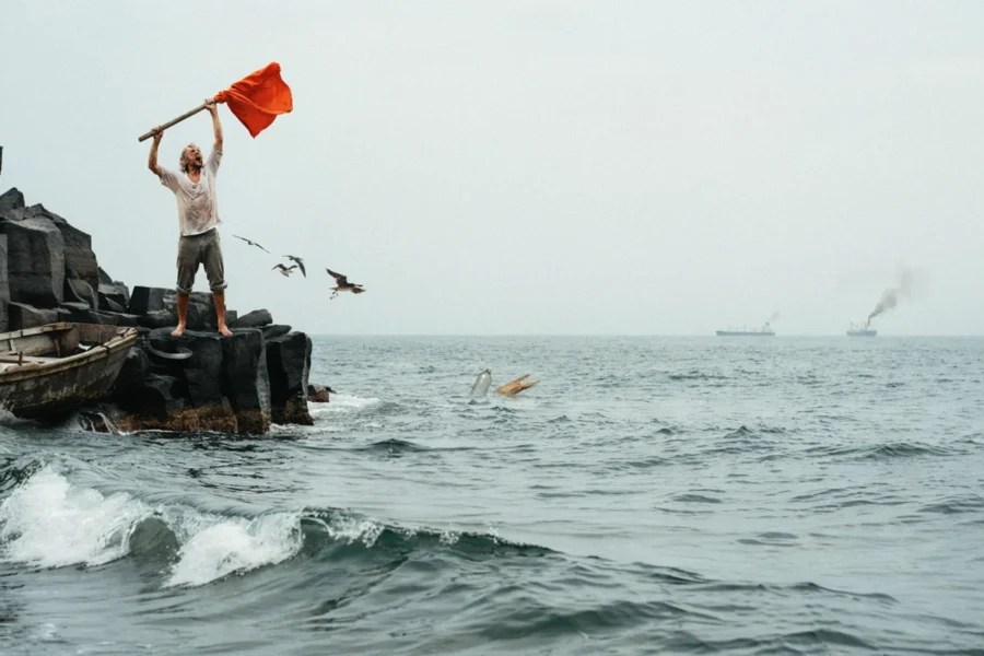 Un homme se dresse sur des structures au bord de l’eau agitant un drapeau rouge vif avec les bras levés au-dessus de vagues grises.