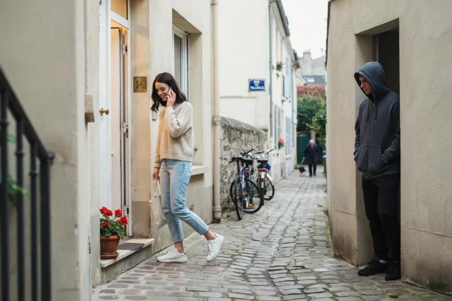 Une femme marche sur un chemin pavé en discutant au téléphone. Un individu vêtu d'une capuche la surveille depuis un passage latéral.